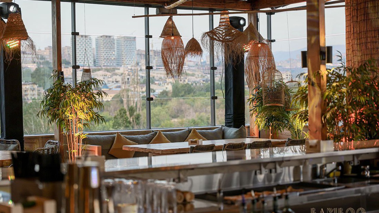 فضای روفتاپ Bamboo Rooftop Bar & Restaurant در ایروان با میزهای چوبی، دکور گرمسیری و چشمانداز شهر با کالسکه