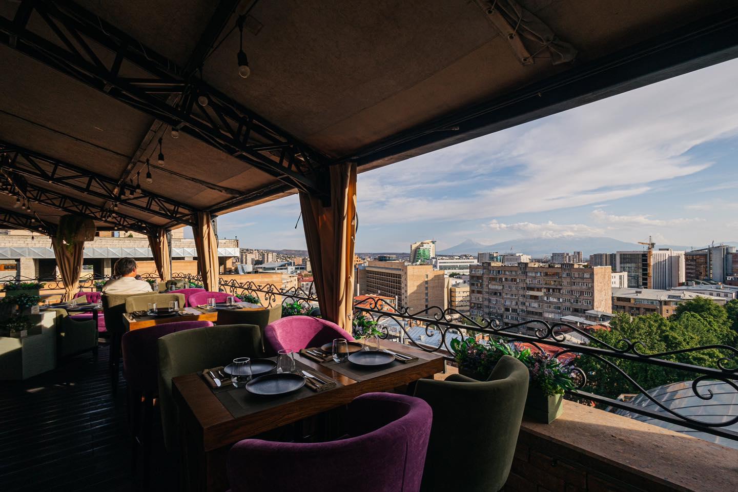 فضای روفتاپ Bamboo Rooftop Bar & Restaurant در ایروان با میزهای چوبی، دکور گرمسیری و چشمانداز شهر با کالسکه