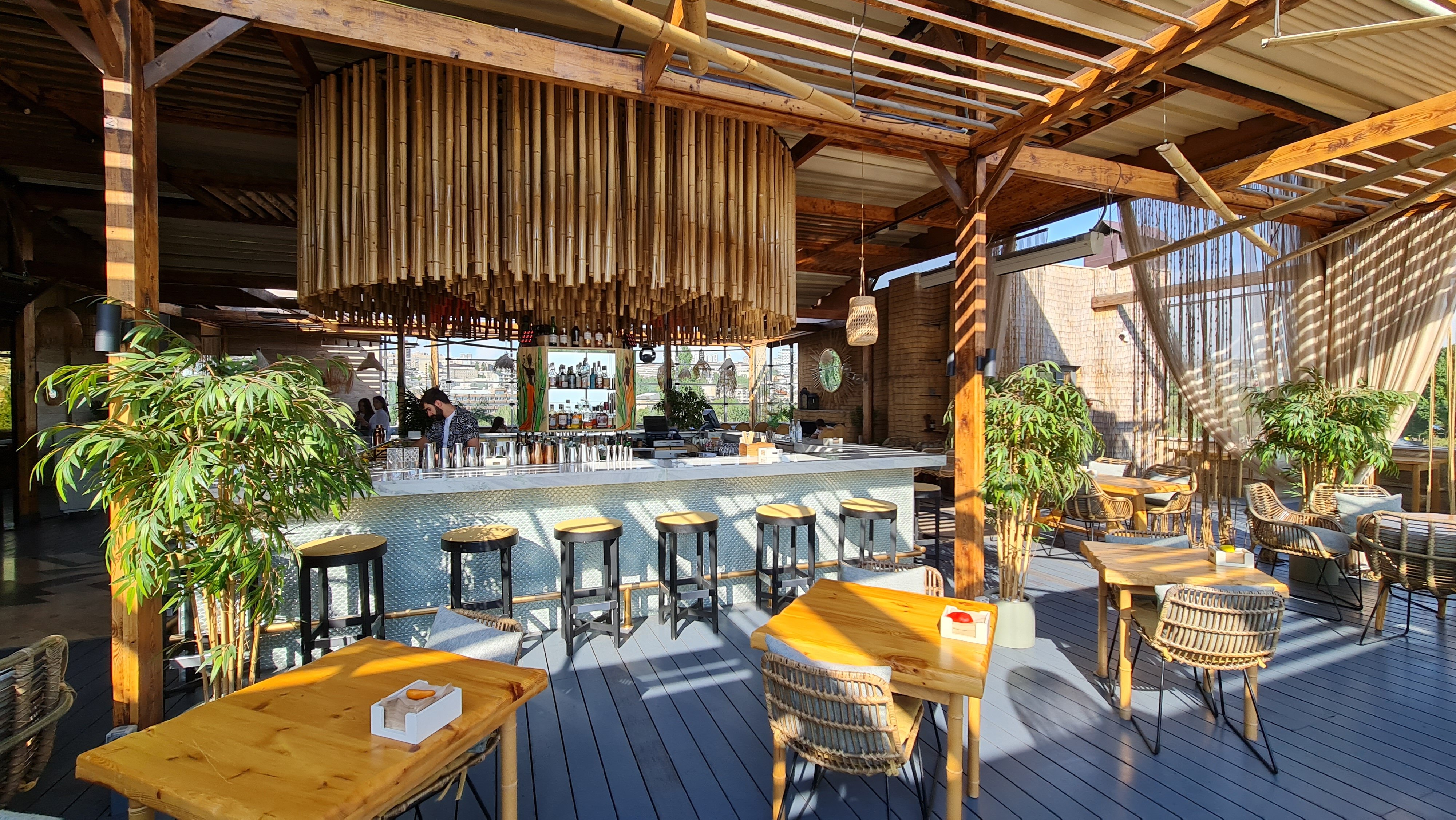 فضای روفتاپ Bamboo Rooftop Bar & Restaurant در ایروان با میزهای چوبی، دکور گرمسیری و چشمانداز شهر با کالسکه